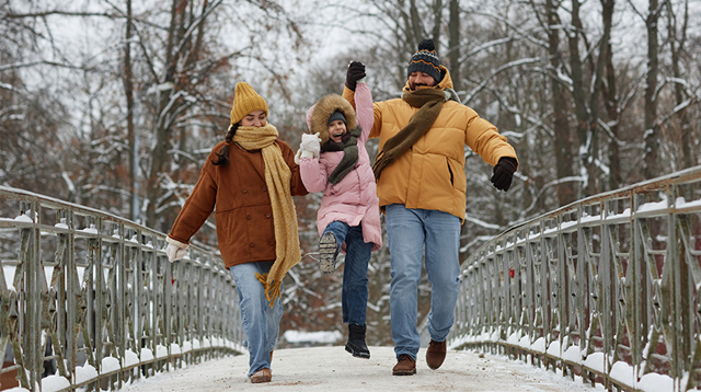 Full length portrait of happy family with daughter jumping up and holding hands while walking towards camera on bridge in winter